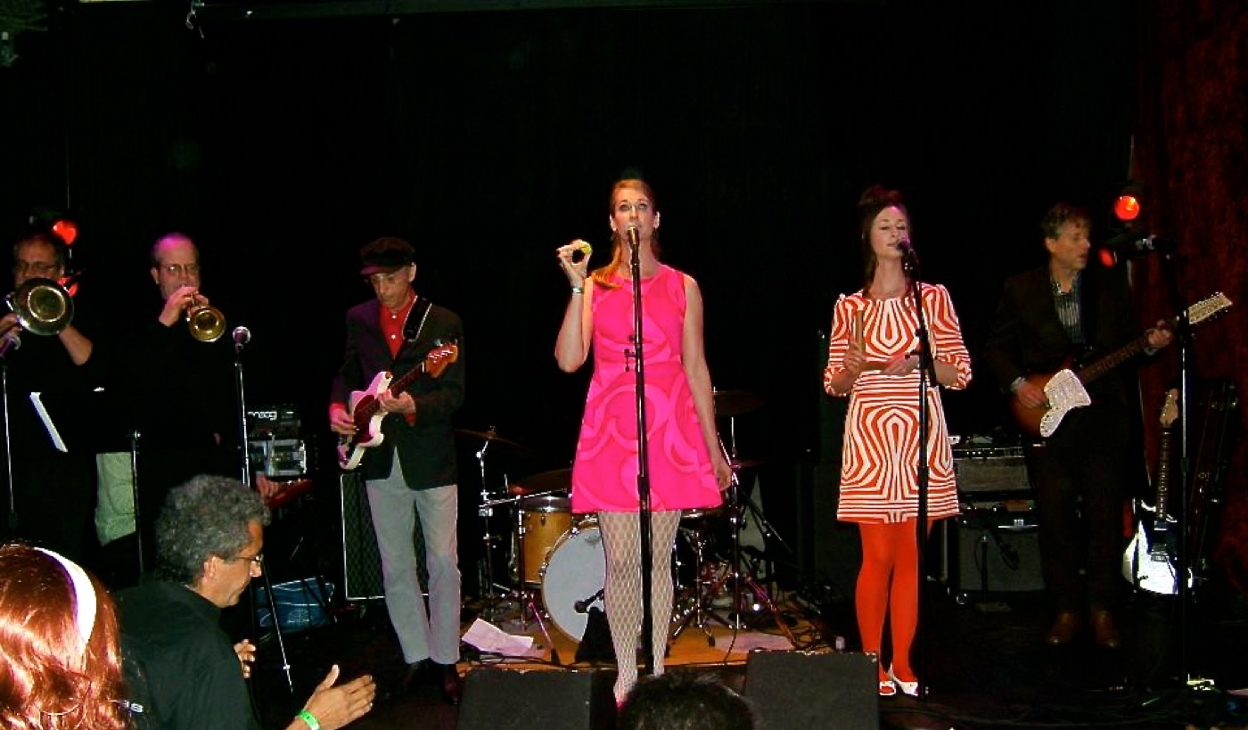 The band onstage with horn section, singers in pink and op-art dresses